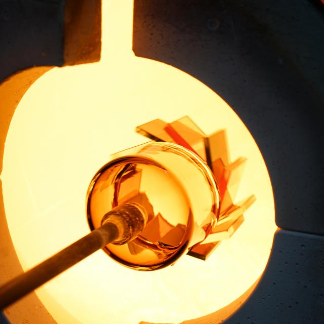 A hot glass piece, still on the end of a blowpipe, is inserted into a furnace to be reheated